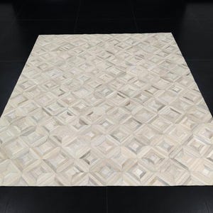 Könnte beinhalten: Ein rechteckiger Teppich mit einem sich wiederholenden geometrischen Muster aus Quadraten und Rauten. Der Teppich besteht aus zahlreichen kleinen, hellen Fellstücken in verschiedenen Nuancen von Cremeweiß und sanftem Grau mit strukturierter Oberfläche.