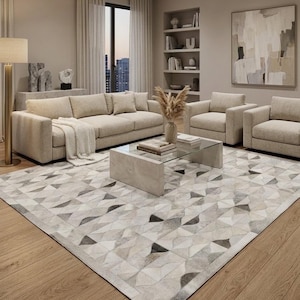 May include: A large area rug featuring a geometric pattern composed of triangular and semi-circular patches in shades of white, gray, and charcoal. The rug has a textured, pieced construction and is laid flat on a light-toned wooden floor.