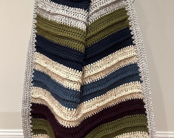 Könnte beinhalten: Eine gestreifte Häkeldecke in den Farben Grau, Grün, Blau und Lila. Die Decke ist in der Mitte gefaltet und wird von einer Person gehalten.