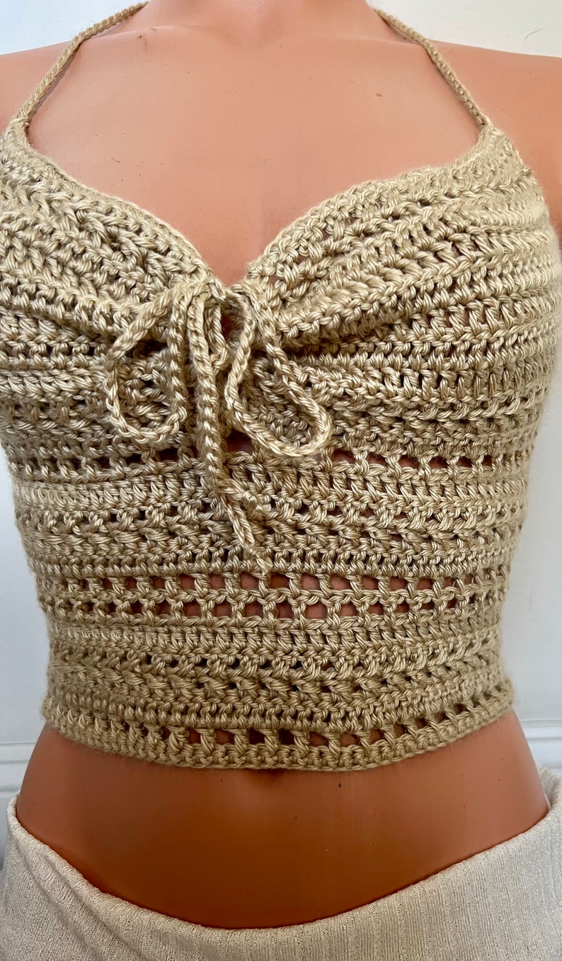 May include: A beige crochet halter top with a tie at the front. The top is made with a delicate open stitch pattern.