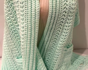 May include: A light green crocheted shawl with fringe. The shawl has a pocket on one side and a V-shaped neckline.