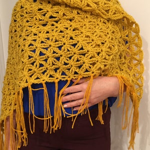 Pu&ograve; includere: Uno scialle all'uncinetto giallo con un motivo a fiore e frange. Lo scialle &egrave; drappeggiato sulle spalle di una persona.