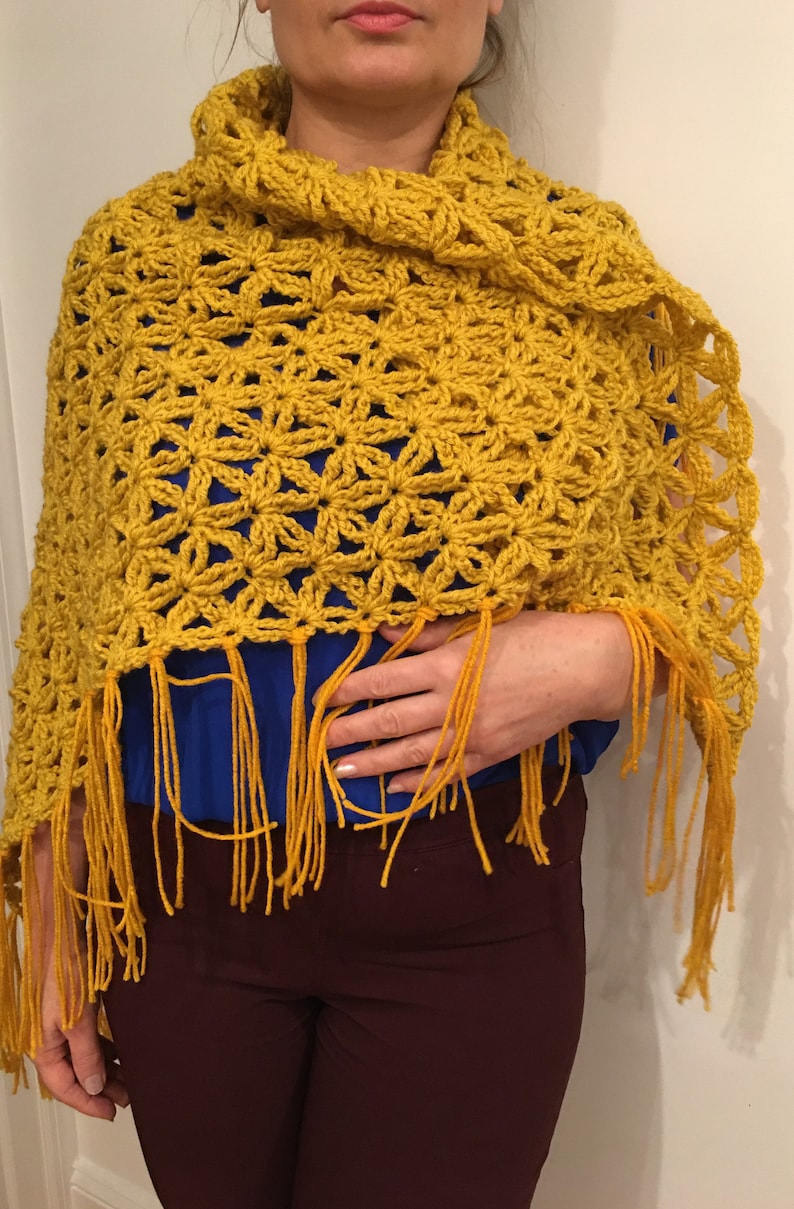 Pu&ograve; includere: Uno scialle all'uncinetto giallo con un motivo a fiore e frange. Lo scialle &egrave; drappeggiato sulle spalle di una persona.