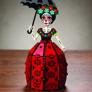 May include: A decorative wooden figurine of a skeleton figure with a painted face, wearing a black top with floral accents and a red skirt featuring black floral and skull cutouts. The figure holds a black umbrella and wears a floral headpiece.