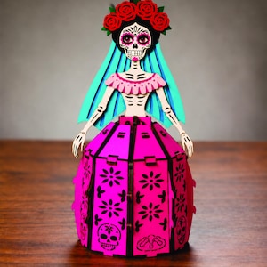 May include: A laser-cut wooden figurine of a skeleton figure wearing a bright pink dress with floral and skull cutouts. The figure has a black skull face with painted details, a crown of red roses, and a flowing turquoise veil over the shoulders.