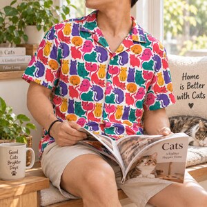 May include: A short-sleeved button-down shirt featuring a repeating pattern of stylized cats in vibrant shades of yellow, orange, pink, purple, red, and teal against a white background, with a classic collar and a front button closure.