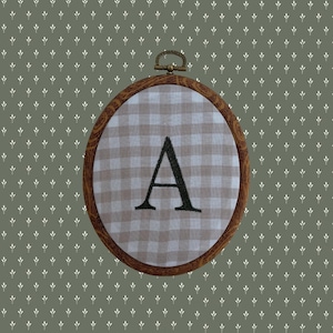 May include: An oval embroidery hoop with a dark wood-toned frame containing light beige and white gingham fabric. A capital letter A is embroidered in black thread in the centre of the fabric. The hoop features a small metal hanging loop at the top.