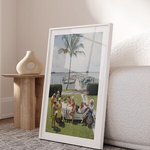 Op de afbeelding: Een ingelijste verticale wandkunstprint met een vintage foto van een groep mensen verzameld rond een witte cabriolet op een grasveld, met een palmboom, een jachthaven en boten op de achtergrond onder een heldere lucht.