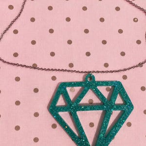 May include: A necklace featuring a large, geometric diamond-shaped pendant made of teal resin with embedded glitter. The pendant is suspended from a thin, metallic chain. The design consists of a hollow, angular frame that mimics the facets of a diamond.