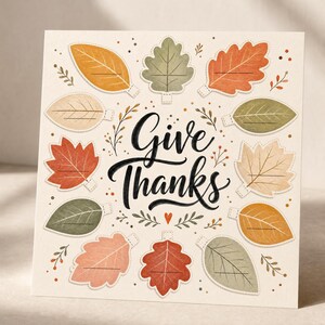Peut inclure: Une carte de vœux carrée sur fond crème avec le texte manuscrit noir Give Thanks au centre. Le motif est entouré de douze feuilles d'automne illustrées dans des tons orange, rouge, vert et beige, chacune comportant une ligne vide.