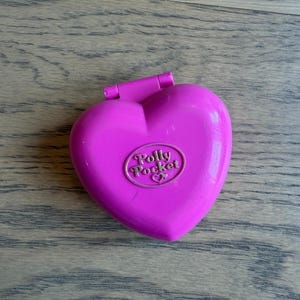 May include: A bright pink heart-shaped Polly Pocket compact case with a hinged lid. The center of the lid features an oval-shaped embossed logo that reads Polly Pocket with a small heart icon underneath the text, all in a matching pink plastic.