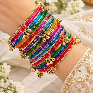 May include: A stack of translucent, multi-colored glass bangles in shades of red, blue, green, yellow, and purple. The bangles feature a textured, faceted surface and are accented with small, dangling gold-toned metal charms.