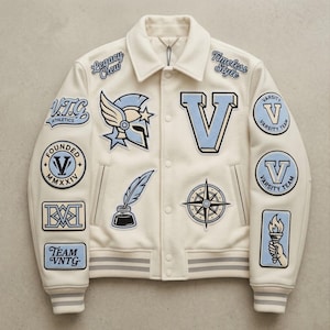 Könnte beinhalten: Eine cremeweiße Collegejacke mit hellblauen und marineblauen Stickaufnähern, darunter ein geflügelter Helm, ein großer Buchstabe V, ein Kompass, eine Feder im Tintenfass sowie der Text Legacy Crew, Timeless Style, V.T.G. Athletics und Team VNTG.
