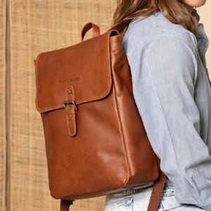 Puede incluir: Una mochila de cuero color canela con solapa plegable, cierre de hebilla única y asa superior. El texto "POUR MARIE" está grabado en la parte frontal de la solapa. La bolsa cuenta con correas ajustables para los hombros y forma rectangular.