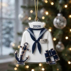 May include: A white and navy blue sailor uniform ornament featuring a navy blue anchor on the left and navy blue binoculars on the right. The collar displays the year 2026 in black text, and a name tag on the right side reads the name Ethan.