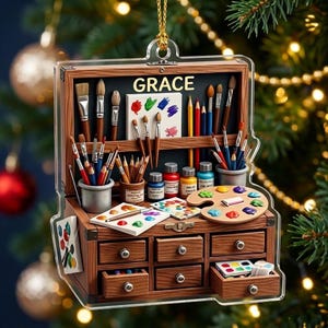 May include: A flat, clear acrylic ornament shaped like an open wooden art supply chest. The interior features the name GRACE, various paintbrushes, colored pencils, paint tubes, and palettes with colorful paint dabs. It has small drawers.