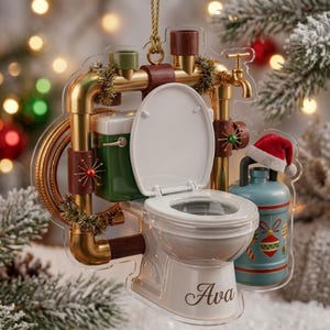 May include: A flat acrylic Christmas ornament featuring a white toilet with the name Ava printed on the base. The toilet is framed by gold-colored pipes, a green tank, a blue water jug wearing a miniature Santa hat, and gold tinsel accents.