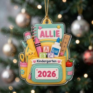 May include: A decorative ornament shaped like a yellow and teal backpack featuring the name ALLIE, the word Kindergarten, and the year 2026. It includes school supplies like a pencil, crayon, glue, ruler, alphabet blocks, and a red apple.