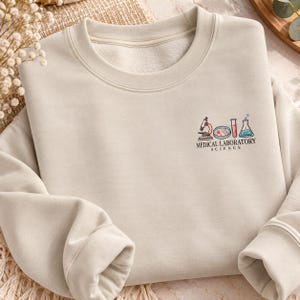 Könnte beinhalten: Ein beigefarbenes Sweatshirt mit Rundhalsausschnitt und einer kleinen Stickerei auf der linken Brust. Das Motiv zeigt ein Mikroskop, eine Petrischale, ein Reagenzglas und einen Erlenmeyerkolben sowie den Text MEDICAL LABORATORY SCIENCE.