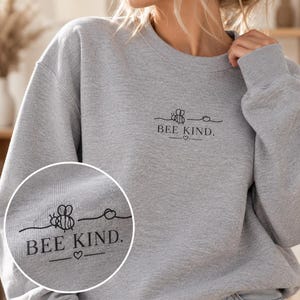 Könnte beinhalten: Ein hellgraues Sweatshirt mit Rundhalsausschnitt und einer schwarzen Strichgrafik auf der linken Brust. Das Design zeigt eine kleine Biene, eine Wellenlinie und den Text BEE KIND. mit einem kleinen Herz zwischen zwei Linien.