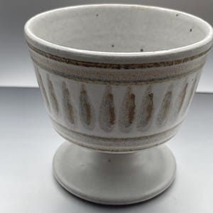May include: A ceramic footed bowl with a speckled off-white glaze. The exterior features a horizontal band near the rim and a series of vertical, teardrop-shaped brown markings around the center, all set against a smooth, light-colored base.