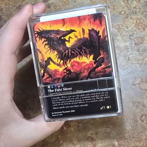 May include: A clear plastic protective case containing a Magic: The Gathering trading card titled The First Sliver. The card features a dark, multi-limbed creature illustration against a fiery orange and red background with black text details.