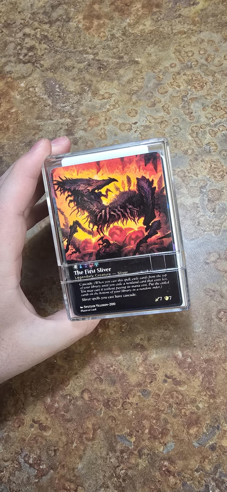 May include: A clear plastic protective case containing a Magic: The Gathering trading card titled The First Sliver. The card features a dark, multi-limbed creature illustration against a fiery orange and red background with black text details.