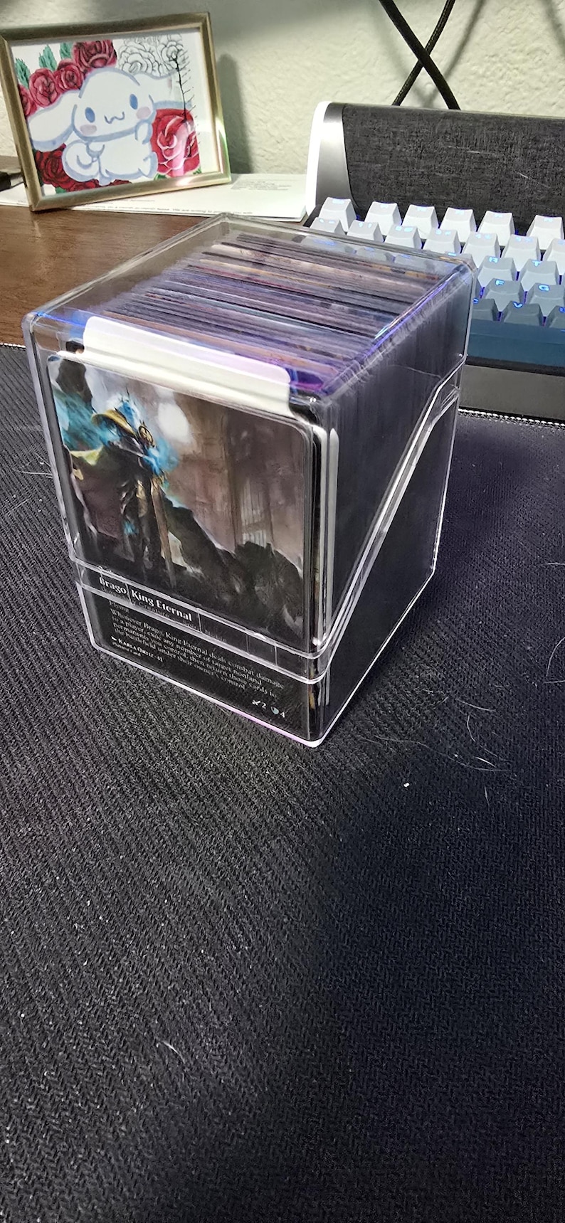 May include: A clear acrylic deck box containing a stack of trading cards. The front card is visible, featuring a fantasy illustration of a character and the text Brago, King Eternal. The box has a hinged lid and a transparent design for storage.