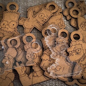 Può includere: Una collezione di ornamenti in legno e acrilico trasparente tagliati al laser con contorni incisi di vari personaggi dei cartoni animati come Bart, Homer e Moe, ciascuno dotato di un foro circolare nella parte superiore per appenderli.