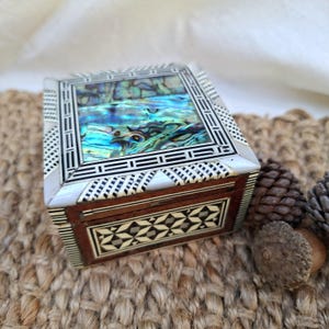 May include: A square wooden box featuring an inlaid lid with a central iridescent abalone shell panel surrounded by black and white geometric patterns. The sides of the box display intricate star-shaped inlays set against dark wood grain.