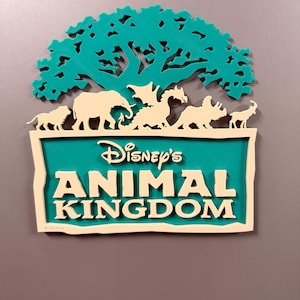 May include: A decorative wall sign featuring the text Disney's ANIMAL KINGDOM in white lettering on a teal background. Above the text is a teal tree silhouette with cream-colored animal figures including a lion, elephant, dragon, and triceratops.