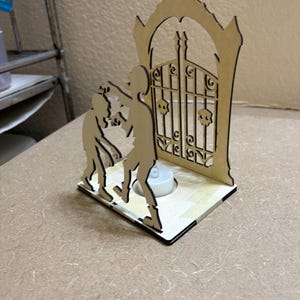 May include: A laser-cut light-colored wooden display featuring two zombie figures standing in front of an ornate arched gate with skull accents. The base includes a circular cutout designed to hold a small white tea light candle.