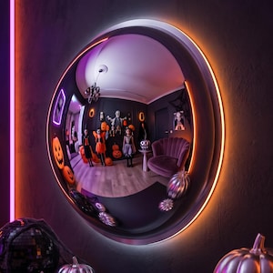 May include: A circular convex wall mirror with a glowing orange LED light ring around its frame. The mirror reflects a room decorated for Halloween, featuring figures in costumes, orange jack-o'-lanterns, and a purple velvet armchair.