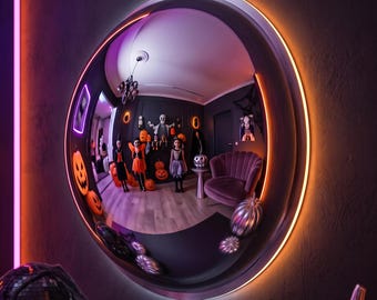 May include: A circular convex wall mirror with a glowing orange LED light ring around its frame. The mirror reflects a room decorated for Halloween, featuring figures in costumes, orange jack-o'-lanterns, and a purple velvet armchair.
