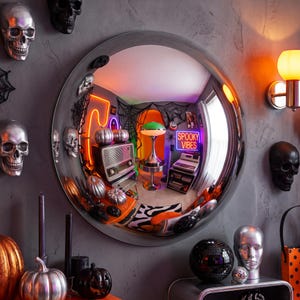 May include: A large circular convex mirror with a silver frame mounted on a gray wall. The mirror reflects a room decorated with Halloween items, including a neon sign that reads SPOOKY VIBES, a green mushroom lamp, pumpkins, and cobwebs.