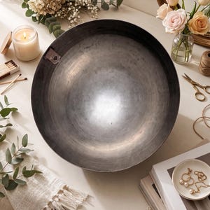 K&ouml;nnte beinhalten: Eine gro&szlig;e, kreisf&ouml;rmige Schale aus dunkelgrauem Metall mit einer glatten, reflektierenden Innenfl&auml;che und einer kleinen Metalllasche am oberen linken Rand. Die Schale hat eine tiefe, beckenartige Form mit einer weiten &Ouml;ffnung.