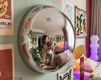 May include: A large, circular, convex wall mirror with a wide, sage green frame. The mirror reflects a room interior, including a person holding a smartphone, a green potted plant, and various colorful wall art pieces and bedding.