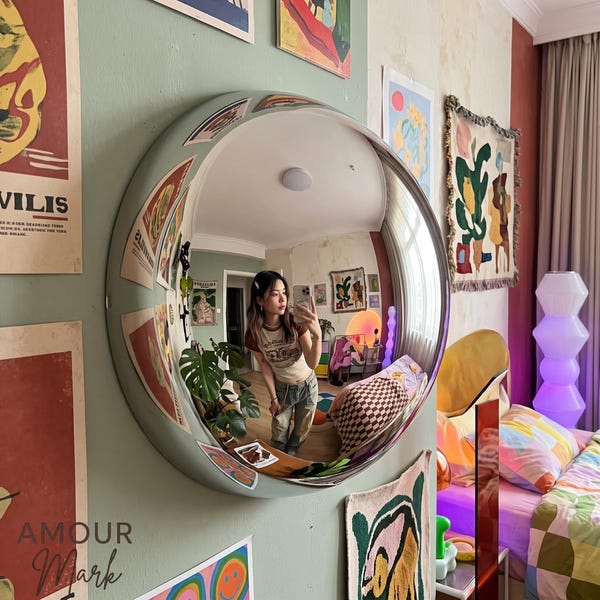 Op de afbeelding: Een grote, ronde, bolle wandspiegel met een brede, saliegroene lijst. De spiegel reflecteert een kamerinterieur, inclusief een persoon die een smartphone vasthoudt, een groene kamerplant en diverse kleurrijke wanddecoraties.