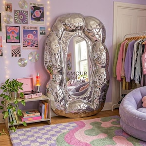 May include: A large, floor-standing mirror with an irregular, organic, and highly reflective silver frame that has a hammered, liquid-like texture. The center of the glass features the black text "AMOUR Mark" printed in a stylized font.