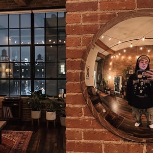 May include: A large circular wall mirror with a dark wood frame mounted on an exposed red brick wall. The mirror reflects a room with wooden floors, string lights, and a person wearing a black hooded sweatshirt and a beanie holding a smartphone.