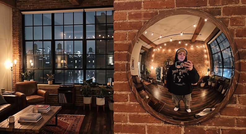 May include: A large circular wall mirror with a dark wood frame mounted on an exposed red brick wall. The mirror reflects a room with wooden floors, string lights, and a person wearing a black hooded sweatshirt and a beanie holding a smartphone.