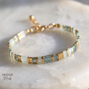 May include: A bracelet featuring alternating rectangular beads in translucent pale green and metallic gold tones. The design includes a gold-toned chain extension with a circular clasp. The text AMOUR Mark is visible in the lower left corner.