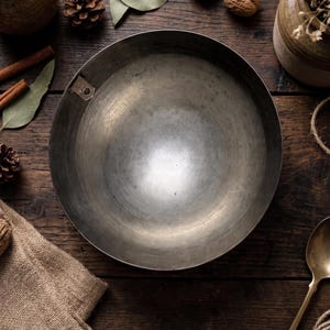 May include: A circular, deep metal bowl with a smooth, brushed gray finish and a single small metal tab attached to the inner rim. The bowl features a wide, open interior and a simple, unadorned design, presenting a functional and utilitarian appearance.