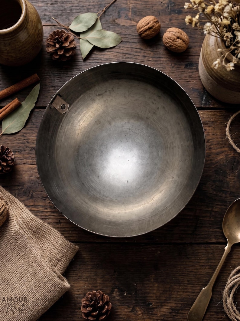 May include: A circular, deep metal bowl with a smooth, brushed gray finish and a single small metal tab attached to the inner rim. The bowl features a wide, open interior and a simple, unadorned design, presenting a functional and utilitarian appearance.