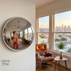 K&ouml;nnte beinhalten: Ein gro&szlig;er, kreisf&ouml;rmiger Wandspiegel mit einem glatten, hellen Rahmen. Der Spiegel reflektiert einen Innenraum mit einer Person in einem rostfarbenen Jumpsuit, einem h&ouml;lzernen Sideboard und einem Teppich. Der Text AMOUR Mark steht unten links.