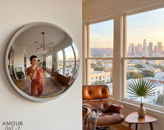 May include: A large, circular wall mirror with a smooth, light-colored frame. The mirror reflects an interior room with a person wearing a rust-colored jumpsuit, a wooden sideboard, and a rug. The text AMOUR Mark is printed in the bottom left corner.