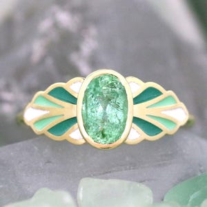 Puede incluir: Un anillo de oro con una piedra preciosa ovalada de color verde claro. El anillo tiene un diseño decorativo con detalles de esmalte blanco y verde azulado. El anillo está sobre una superficie gris con piedras verdes en el fondo.