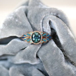 Puede incluir: Un anillo de oro rosa con una piedra ovalada turquesa rodeada de un dise&ntilde;o de esmalte azul y dorado.