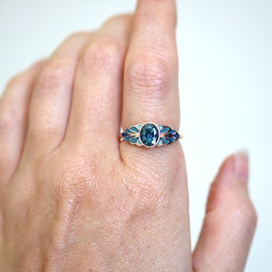 Puede incluir: Un anillo de oro con una piedra ovalada azul rodeada de esmalte azul y blanco. El anillo est&aacute; puesto en un dedo.