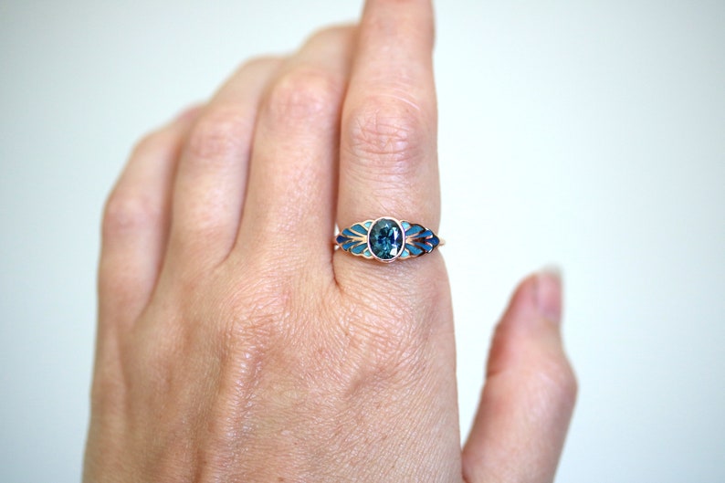 Puede incluir: Un anillo de oro con una piedra ovalada azul rodeada de esmalte azul y blanco. El anillo est&aacute; puesto en un dedo.